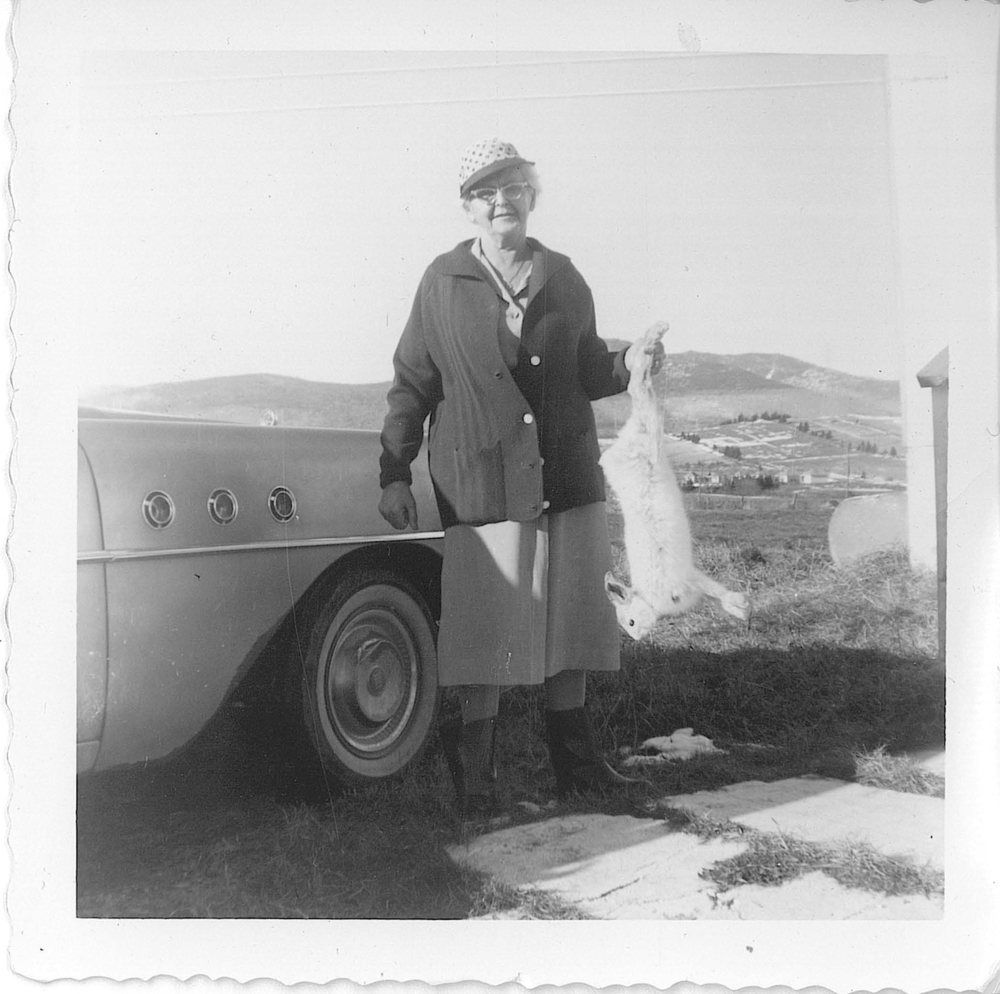 Photographie d’archives en noir et blanc montrant Rose Lachance, âgée, portant une casquette, un tricot, une jupe et des bottes de pluie. Elle tient un lièvre mort dans sa main gauche. Derrière elle se trouve un véhicule. En arrière-plan, on aperçoit des champs et des montagnes.