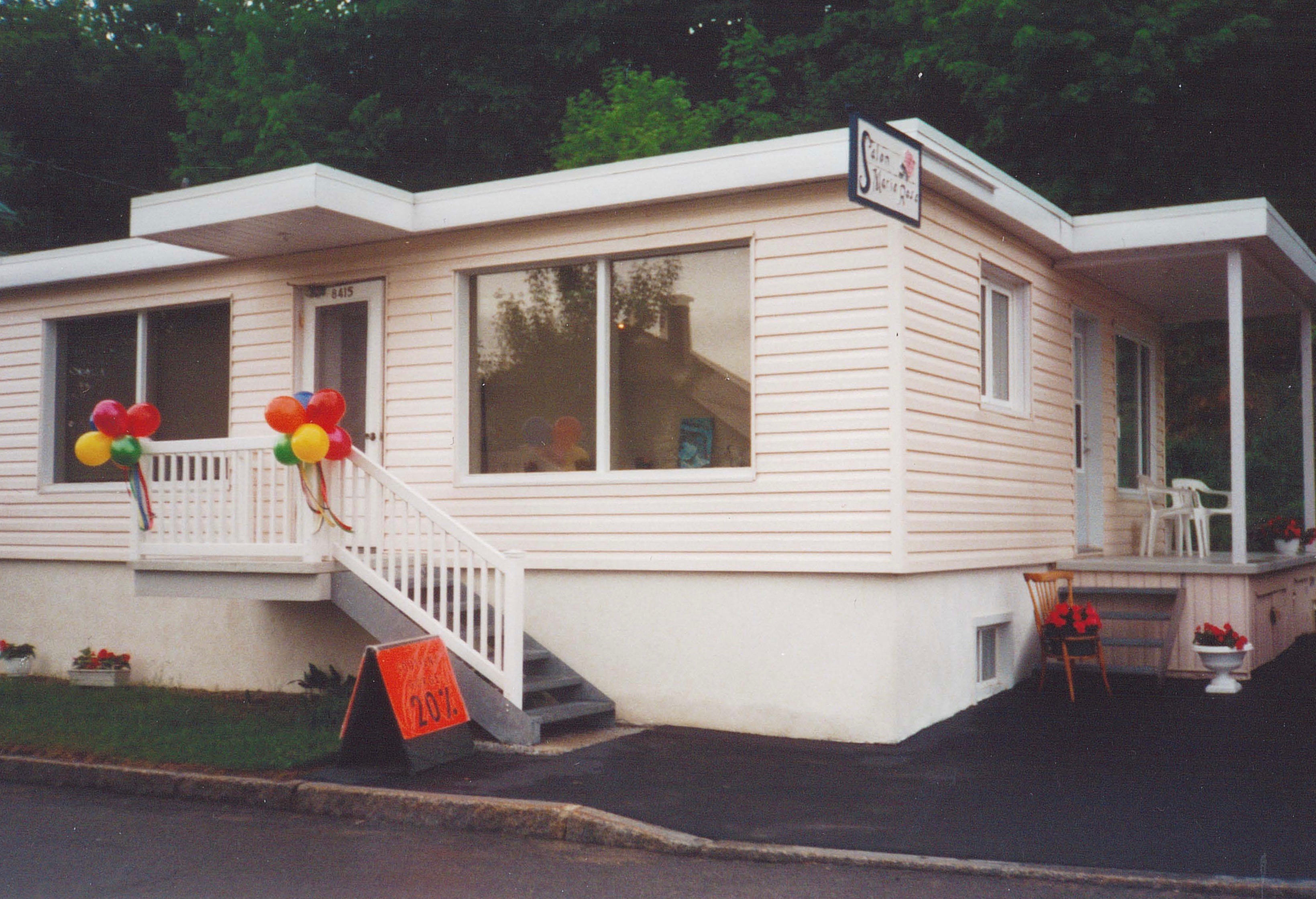 Photographie couleur d’une maison rose pâle à un étage. La vue de trois-quarts montre deux portes d’entrée, une sur le côté et une à l’avant. Sur le côté de la maison, on voit des chaises blanches sur la petite galerie et des fleurs rouges en pot en/au bas des escaliers. À l’avant, des ballons rouges, jaunes, verts et bleus sont accrochés à la balustrade de la galerie. À côté de l’escalier situé à l’avant, une affiche orangée indique « Soutien-gorge - 20% ». Une enseigne où on lit « Salon Marie-Rose » est accrochée perpendiculairement à la façade, de façon à être visible de la rue.