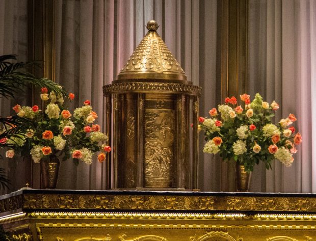 Photographie couleur d’un tabernacle cylindrique en métal repoussé richement travaillé qui est déposé sur un autel entre deux grands vases contenant des bouquets de fleurs roses et blanches. Le haut du tabernacle est de forme conique.