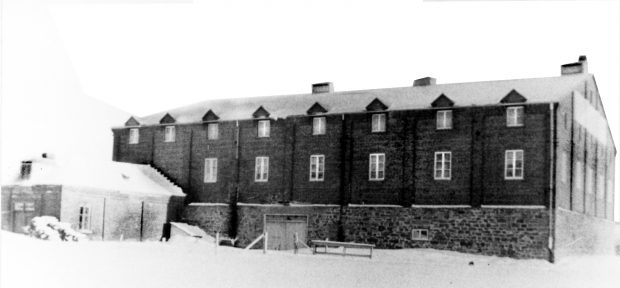 Photographie d’archives en noir et blanc montrant la fabrique de vins A. Toussaint et Cie. Il s’agit d’un grand bâtiment en briques à deux étages. Le mur du bâtiment présente/est percé de deux rangées de huit fenêtres.