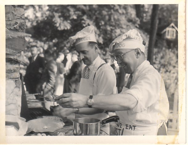 Image en noir et blanc de 2 hommes au barbecue, portant des toques et tabliers de chef ludiques ; chacun des deux porte aussi une chemises et une cravate.