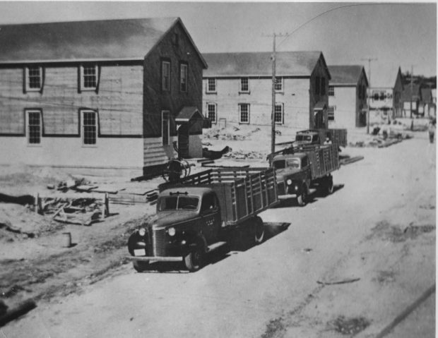 Photo en noir et blanc de deux camions stationnés sur la route à l'extérieur d'un casernement. Plusieurs bâtiments de caserne sont visibles sur le côté gauche de la route.