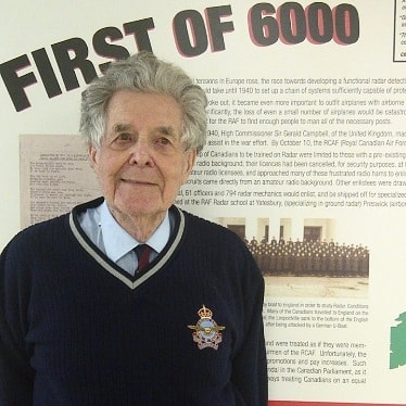 Un homme plus âgé aux cheveux gris portant un chandail bleu et souriant. Il se tient devant un panneau de texte intitulé « Le premier de 6000 ».