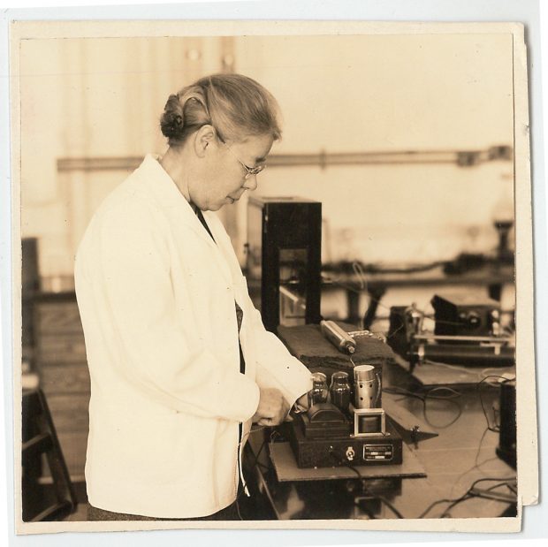 Image en tons sépia d'une femme âgée, portant un sarrau blanc et examinant une petite machine équipée de tubes à vide.