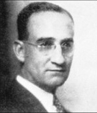 Portrait en noir et blanc granuleux d'un homme blanc portant des lunettes et des cheveux lissés vers l’arrière.