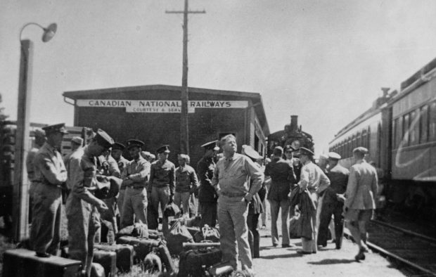 Soldats debout sur un quai de gare. Locomotive et wagons à droite. Derrière eux se trouve la gare avec une pancarte indiquant : « Société de chemins de fer Canadian National ; Courtoisie et service ».