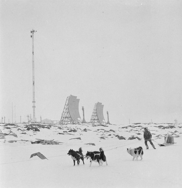 Photographie en noir et blanc d’une personne avec trois chiens tirant un traîneau dans l’Arctique. Une tour radio et des antennes radar sont en arrière-plan.