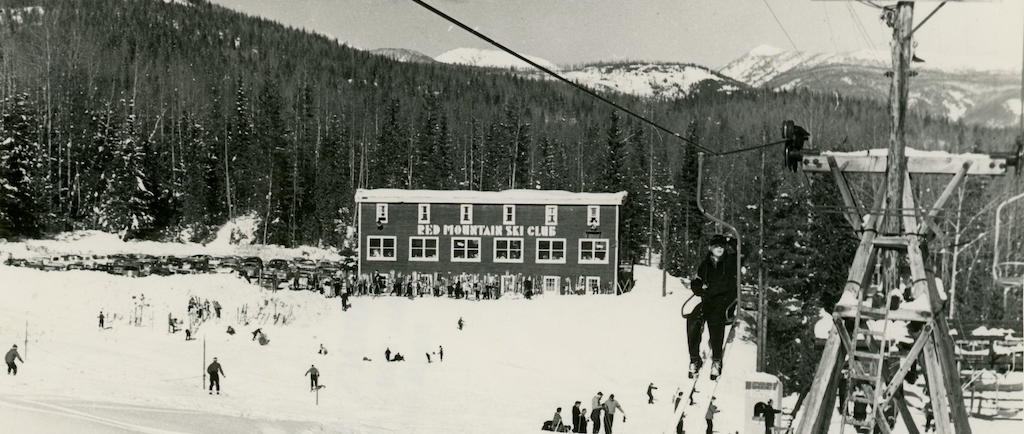 Photo en noir et blanc d’un skieur sur un télésiège. Au loin, on voit le chalet du Club de Ski de Red Mountain.