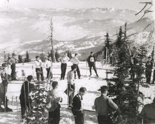 Photo en noir et blanc de plusieurs skieurs hommes portant leur dossard de course debout en haut d’une montagne. Une vaste chaîne montagneuse est visible en arrière-plan.