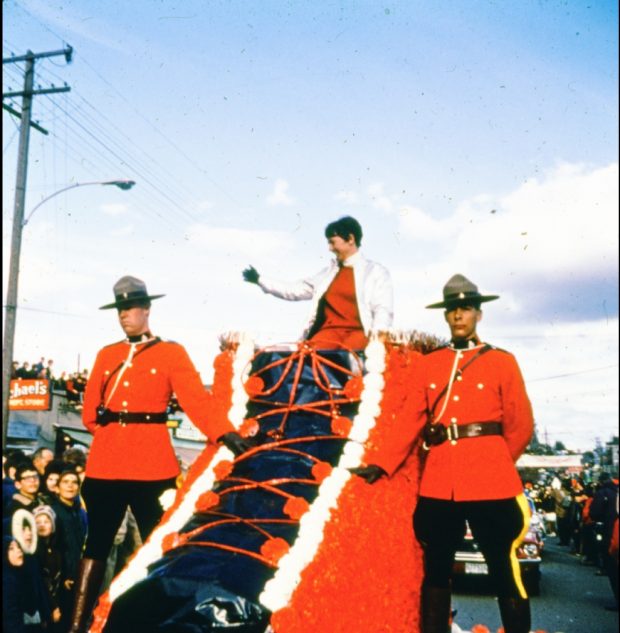 Nancy Greene est assise sur un char de parade en forme de botte de ski rouge, saluant la foule avec deux officiers de la Gendarmerie Royale qui marchent à côté du char. 