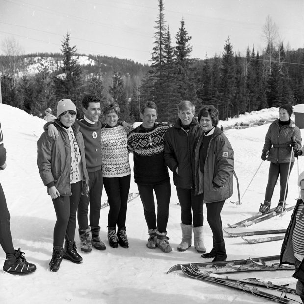 Photo en noir et blanc d’un groupe de six personnes, incluant trois hommes et trois femmes sur la montagne de ski entourées d’équipement de ski. 