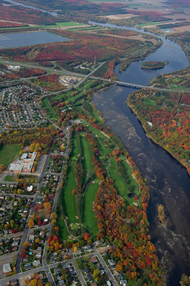 Photographie en couleurs démontrant une vue aérienne du parcours de golf en automne, aménagé le long de la rivière Saint-François. On y voit aussi le développement de la ville et de la flore autour du parcours.