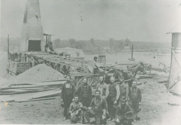 Photographie en noir et blanc de plusieurs hommes devant un haut fourneau en pierre et en brique des forges McDougall sur la rive de la rivière Saint-François.