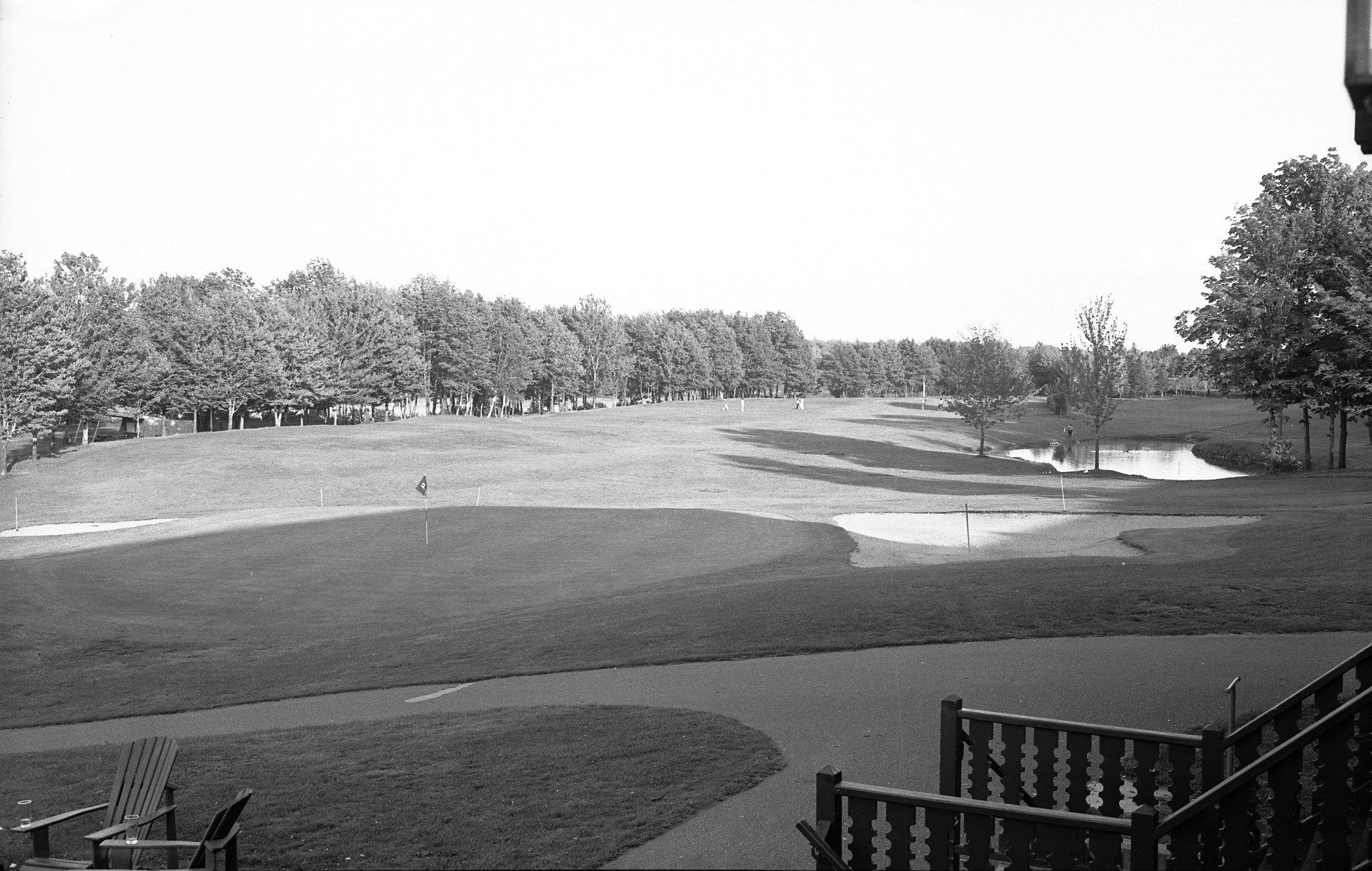 Photographie en noir et blanc démontrant une vue du perron du chalet du parcours de golf. On y voit des drapeaux de trous de golf et un espace de repos pour les golfeurs.