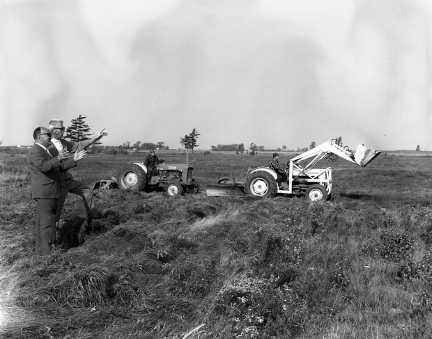 Photographie en noir et blanc de deux hommes supervisant le début des travaux du terrain de golf sur un terrain vague. Derrière eux, deux hommes manœuvrent de la machinerie.
