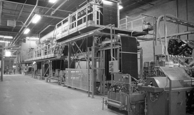 Photographie en noir et blanc de l’intérieur de l’usine Soprema. On y voit plusieurs machines dans un local en béton.