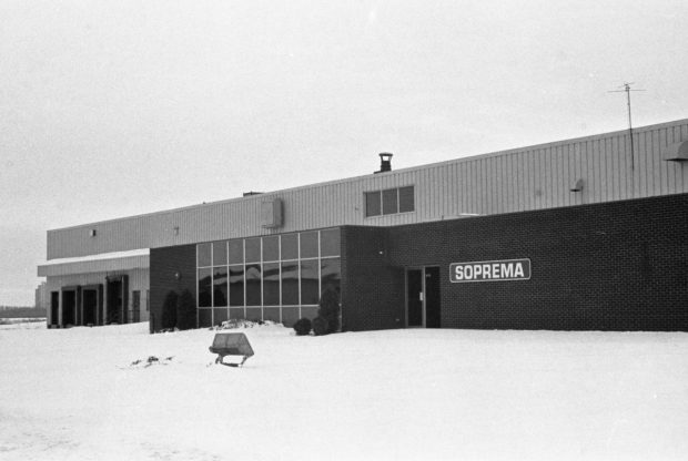 Photographie en noir et blanc de la façade de l’usine Soprema. On y voit l’enseigne sur la grande baie vitrée et trois quais de chargement.