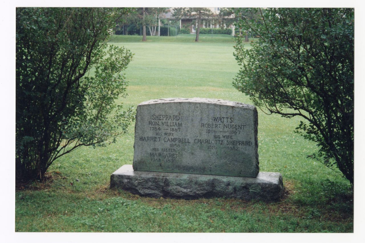 Photographie en couleurs d’une pierre tombale rectangulaire entre deux arbustes. Cinq noms y sont inscrits, notamment Robert Nugent Watts, Charlotte Sheppard, William Sheppard et Harriet Campbell.