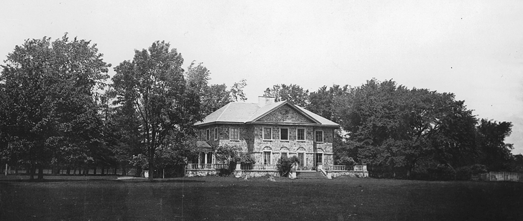 Photographie en noir et blanc du Grantham Hall, une grande maison en pierre de deux étages, entourée d’un perron en pierre, d’arbres et d’arbustes.