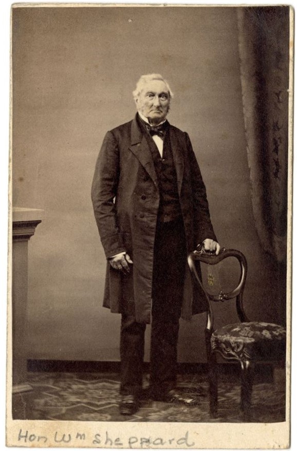 Photographie d’un homme debout derrière une chaise, vêtu d’un long veston noir.