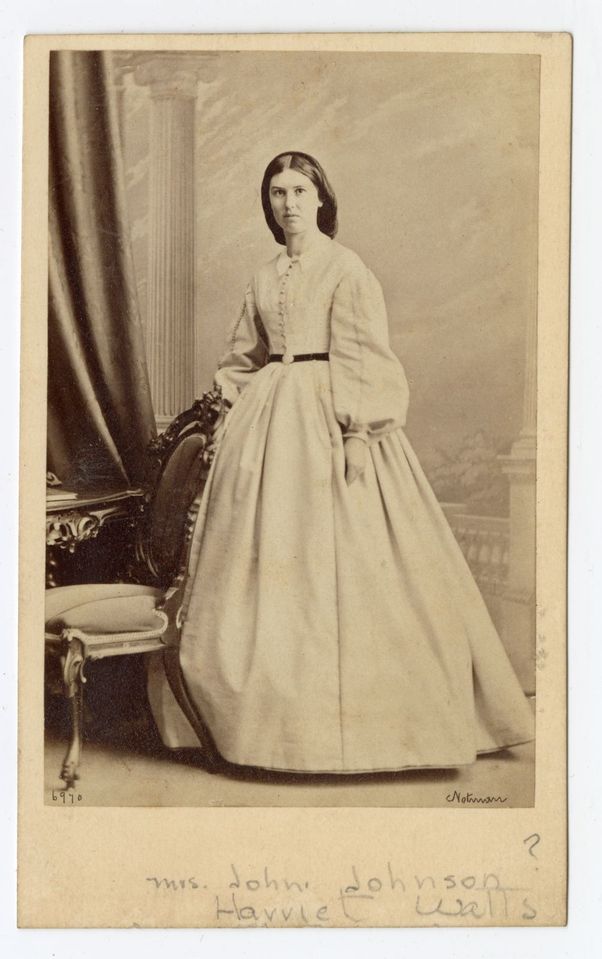 Photographie en noir et blanc d’une femme debout derrière la chaise d’un bureau en bois sculpté, portant une longue robe aux manches bouffantes et un filet dans ses cheveux.