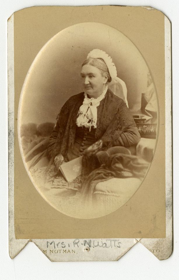 Photographie d’une femme posant assise, un livre sur ses genoux. Elle porte un châle en dentelle, un foulard au cou et un chapeau.