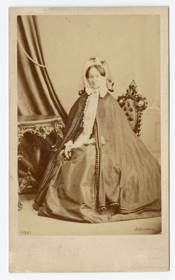 Photographie en noir et blanc d’une femme assise à un bureau en bois sculpté, vêtue de longs vêtements décorés sur les bordures et coiffée d’un chapeau.