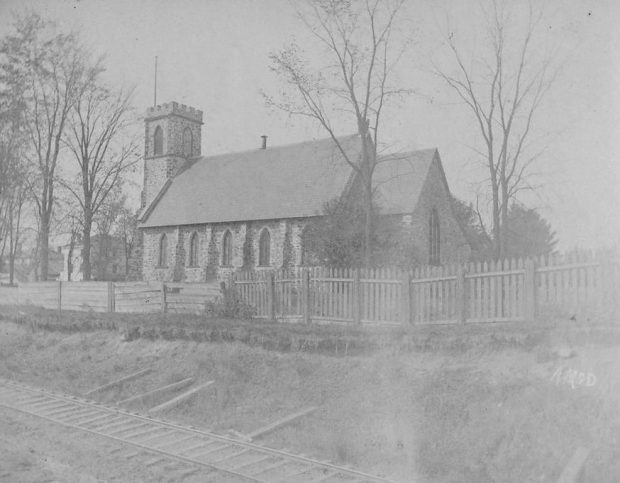 Photographie en noir et blanc de l’église anglicane St. George. Il s’agit d’une église en pierre avec de grands vitraux et un clocher imposant à l’avant. Elle est entourée de quelques arbres et se trouve en bordure du chemin de fer.