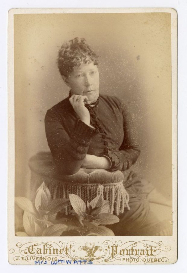 Photographie en noir et blanc d’une femme posant assise, s’appuyant sur un coussin, vêtue d’une longue robe aux manches décorées par des boutons.