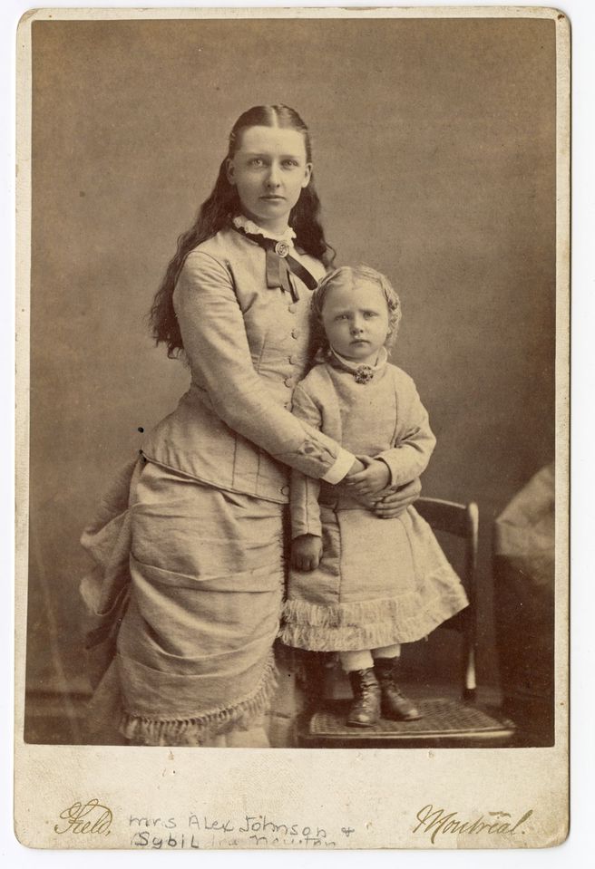 Photographie en noir et blanc d’une femme et d’une petite fille, posant debout, vêtues de longues robes décorées par un ruban et ornées d’une broche au cou. L’enfant est debout sur une chaise pour être au niveau de sa mère.