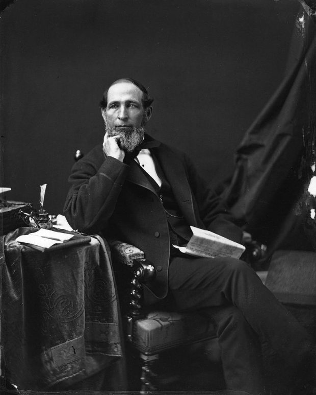 Photographie en noir et blanc d’un homme assis sur un fauteuil avec un journal à la main.