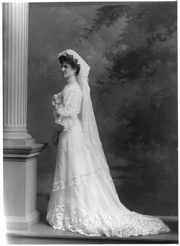 Photographie en noir et blanc d’une femme posant près d’une colonne, vêtue de sa robe de mariée avec de la dentelle et coiffée d’un voile.