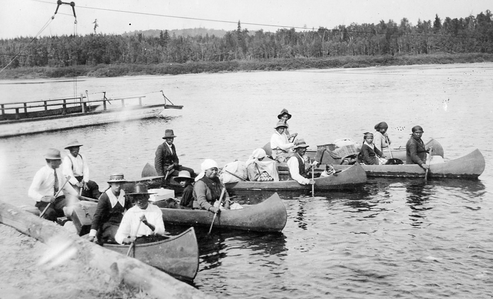 Sur la rivière, des Autochtones sont installés dans cinq canots remplis de bagages.