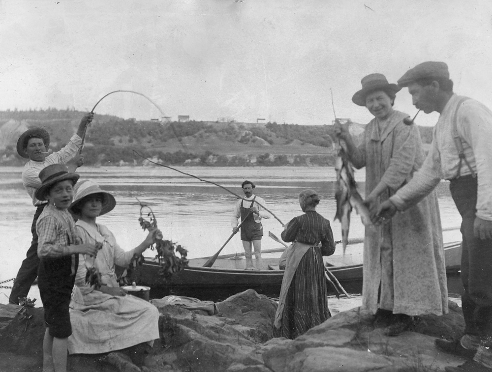 Une famille pêche sur les berges du Saint-Maurice. Un jeune garçon tient une canne à pêche et une femme exhibe plusieurs poissons.