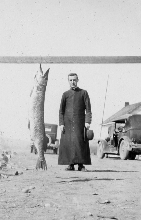 Un abbé pose à côté d'un gros brochet suspendu à une poutrelle.