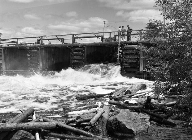 Trois hommes se tiennent sur un barrage fait de rondins de bois. Une vanne du barrage est ouverte et laisse passer un torrent d'eau et de billots.