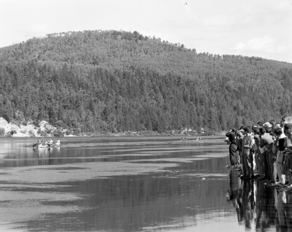 Sur la berge, les spectateurs s'approchent le plus près possible de l'eau pour voir passer les canotiers qui semblent minuscules au milieu de la large rivière et des hautes falaises.