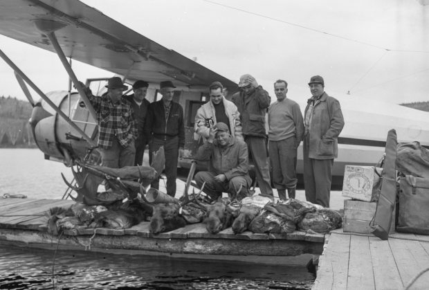 Huit hommes posent sur un quai devant un hydravion avec quatre carcasses d'orignaux.