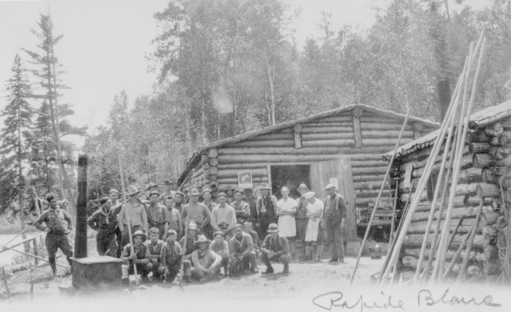 Un groupe de travailleurs posent devant de petites cabanes en bois rond en pleine forêt.