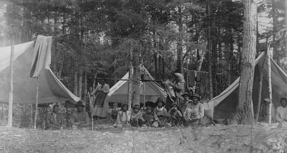 Des Autochtones de tous âges posent au milieu de tentes rudimentaires entourées de hauts conifères.