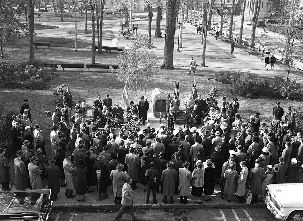 Une foule est rassemblée près d’un parc. Les gens sont regroupés devant un grand rocher sur lequel est posé une plaque de bronze. Un homme s’adresse à eux devant des micros.
