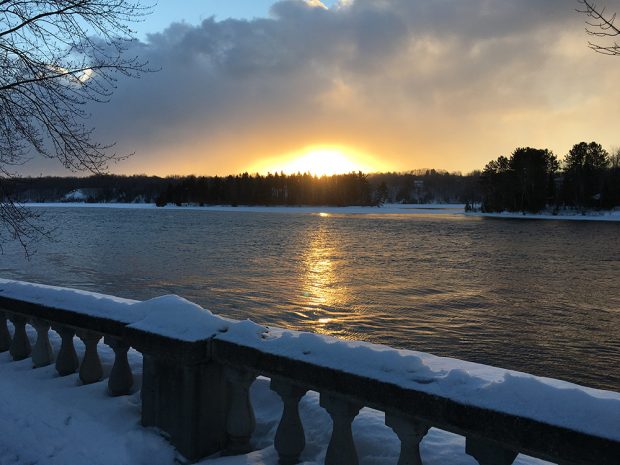  Le coucher de soleil se reflète sur l’eau de la rivière. Une balustrade enneigée occupe le premier plan.