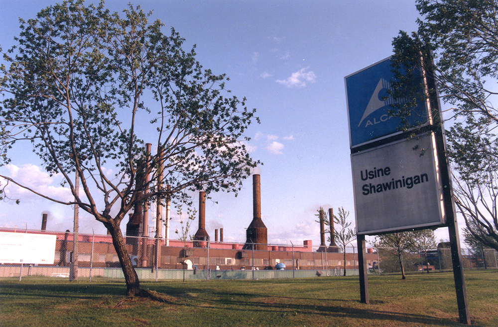 Le premier plan est occupé par un pancarte indiquant : Alan, Usine Shawinigan. En arrière plan, derrière une clotûre, l’usine et ses cheminées dominent le paysage.