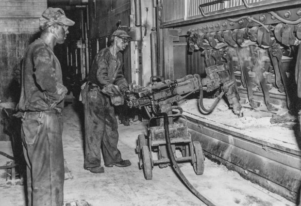 Deux employés portant des vêtements de laine, des casquettes et des petites lunettes manipulent de la machinerie près d'une cuve d'aluminium.