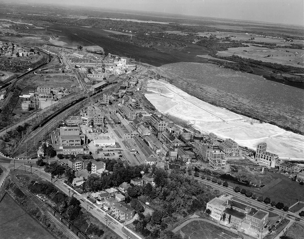 Vue aérienne du l'immense site de l'usine qui s'étend près de la rivière sur laquelle flotte à la fois des milliers de billots de bois et des résidus de chaux blanchâtres.