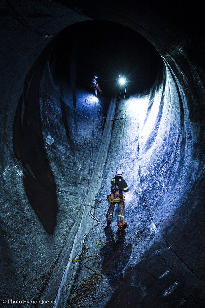 Un travailleur muni d’un équipement d’escalade se tient dans la lumière d’un projecteur à l’intérieur d’un immense tunnel de béton qui fait office de conduite forcée.