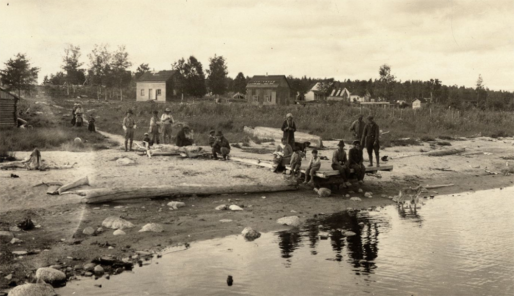 Un groupe de personnes se tient sur une plage. Derrière eux, de modestes maisons en bois sont dispersées. 