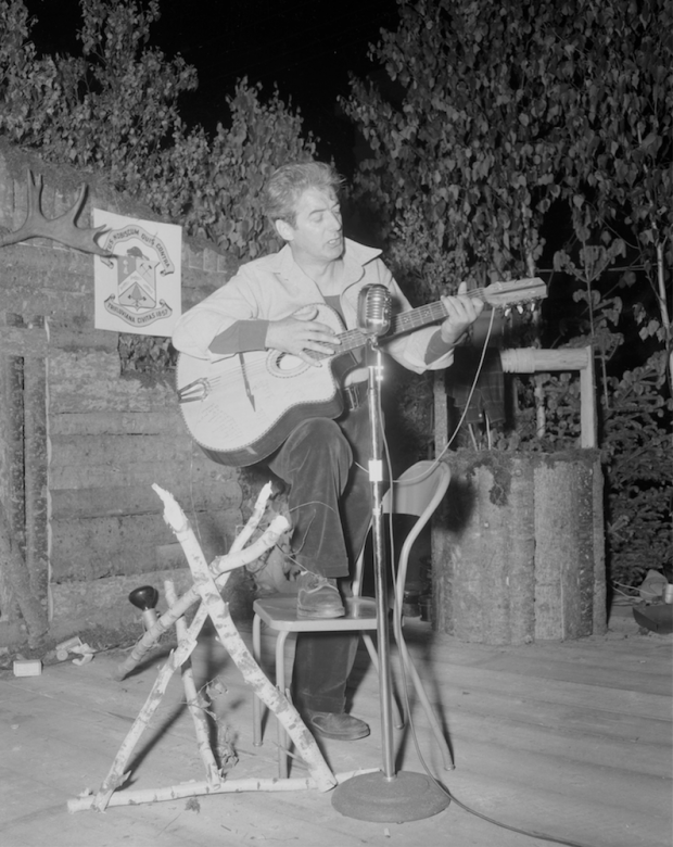 Félix Leclerc chante et joue de la guitare sur une scène décorée de bois rond