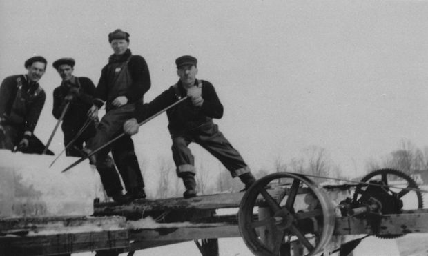 Photographie en noir et blanc de quatre coupeurs de glace chargeant la remorque d’un camion de gros blocs de glace. 