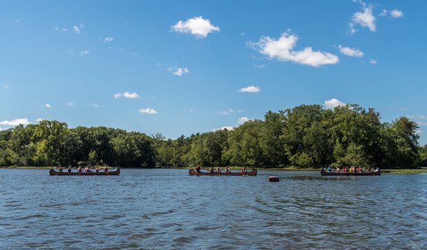 Photographie en couleur de trois canots un à la suite de l’autre sur une rivière. Les canots sont remplis de personnes. Sur la rive, on voit un boisé verdoyant.